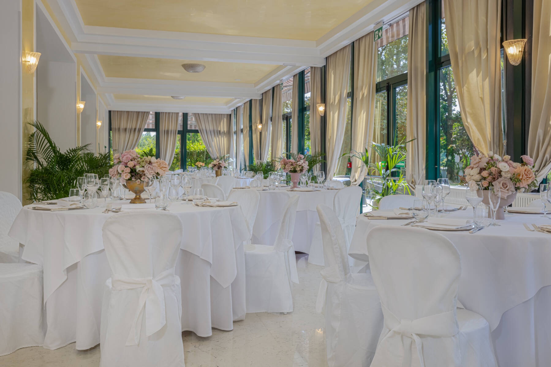 A veranda laid for a wedding, pastel florals and dressed chairs beneath a soft golden ceiling, the park beyond the glass, Villa Pace Park Hotel Bolognese