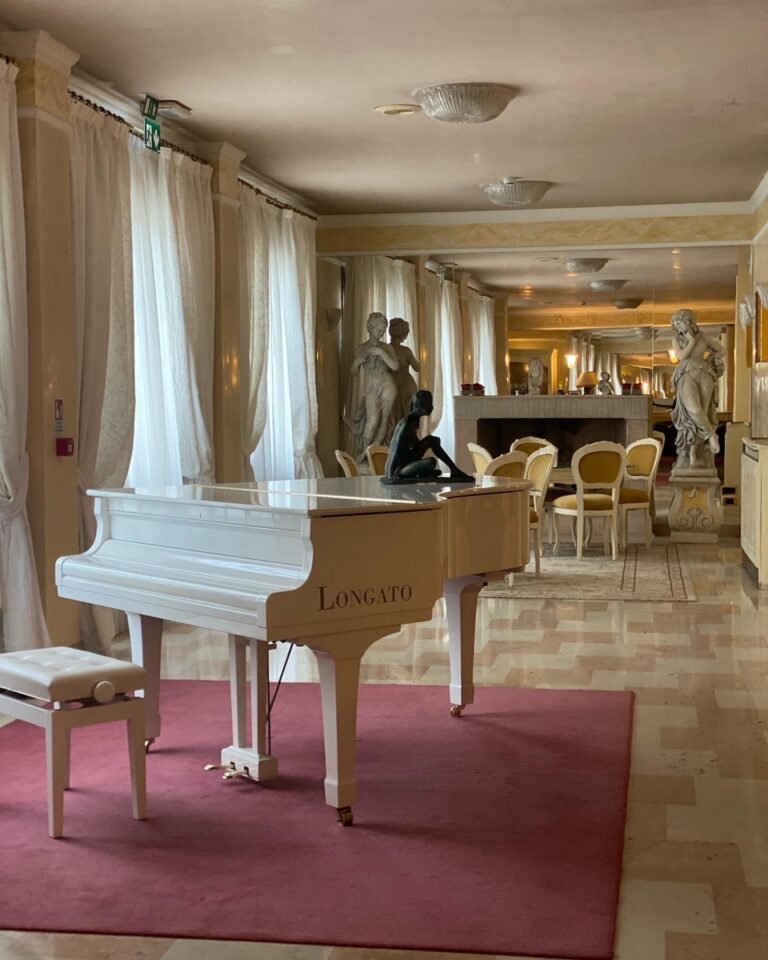 A white Longato grand piano on a rose-coloured rug, marble statues and salon light behind, Villa Pace Park Hotel Bolognese