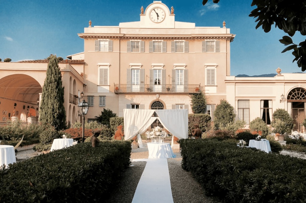 A white-draped ceremony arch at the end of a pale aisle between clipped hedges, with the villa behind