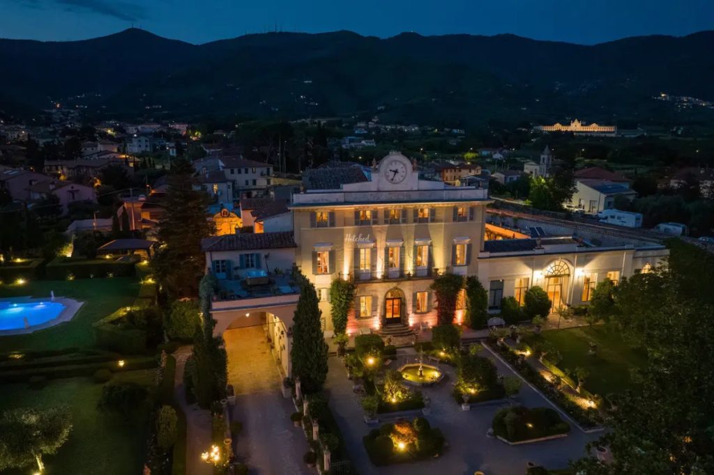 The villa and its lit gardens at dusk, with hills behind