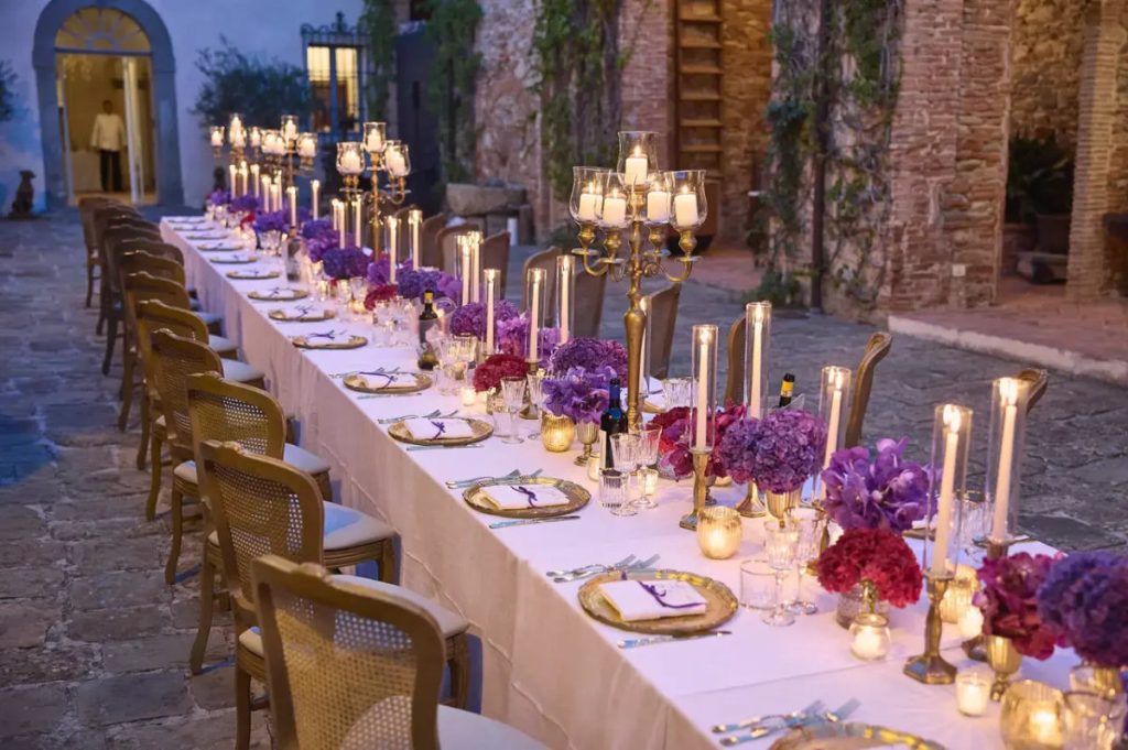 A long table with candles and purple flowers on a stone terrace at dusk