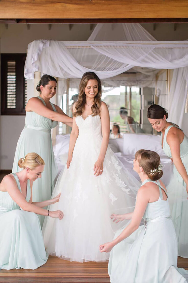 Bridesmaids helping the bride with her dress beside a canopy bed, Villa Susanna St Lucia