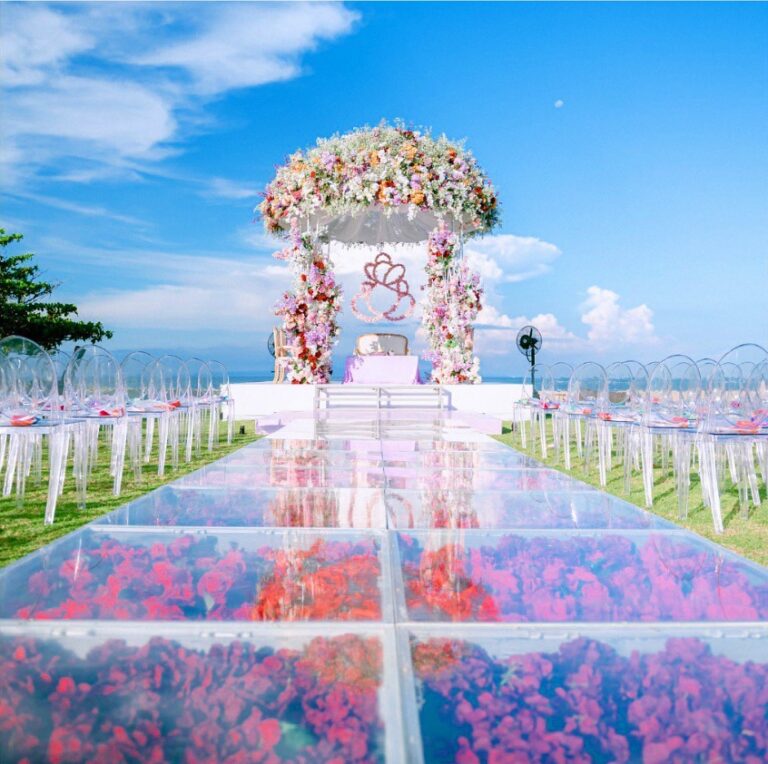 Floral mandap at the end of a glass aisle over pink flowers, with sea and sky behind, Vivaah Weddings