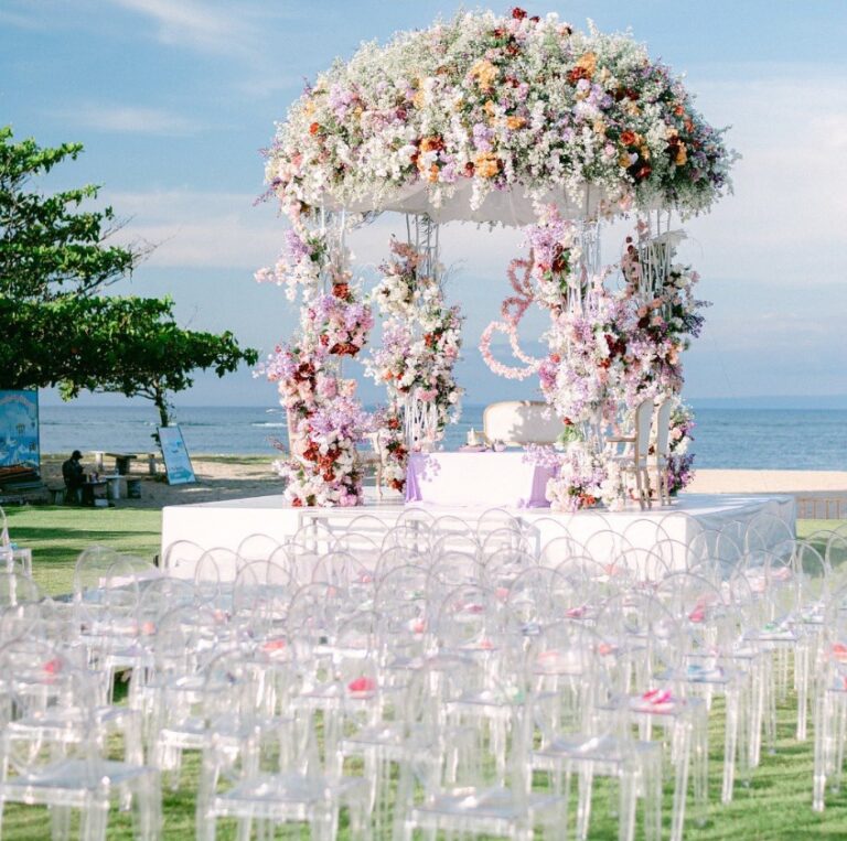 Round floral canopy beside the sea with rows of clear ghost chairs, Vivaah Weddings