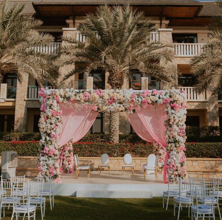 Pink and white floral mandap with draped curtains among palms, with white chairs, Vivaah Weddings