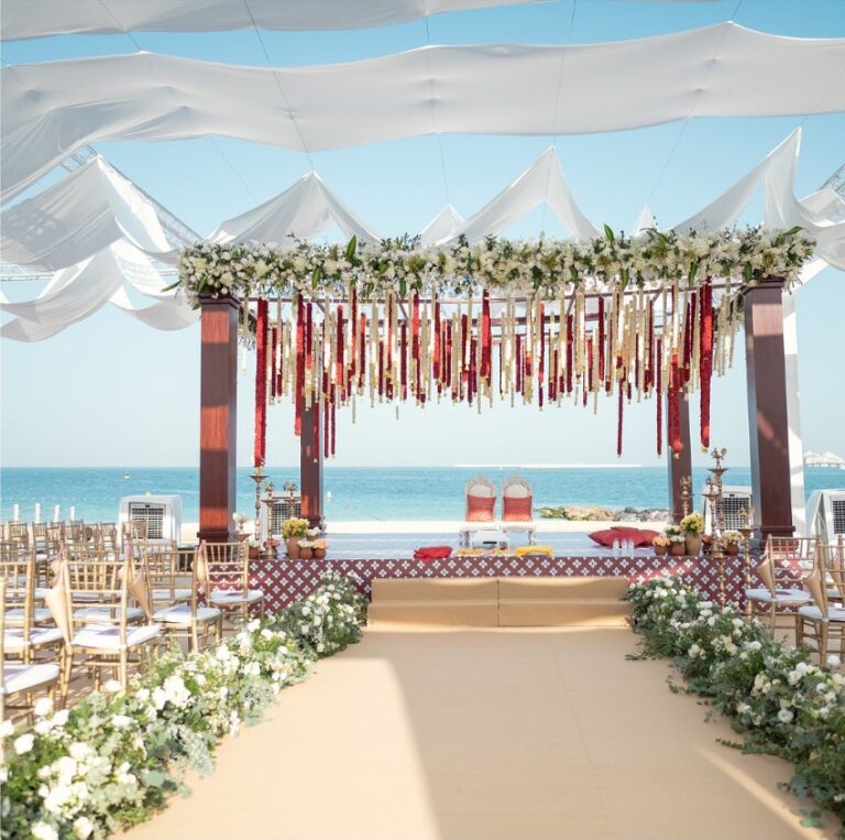 Beachfront mandap hung with red and cream tassels at the end of a flower-lined aisle, Vivaah Weddings