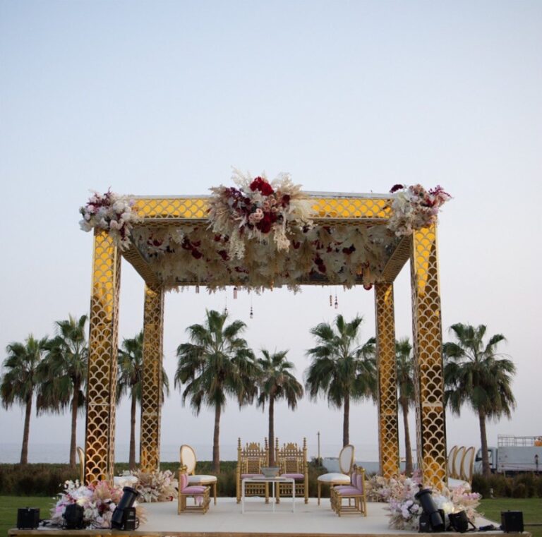 Gold lattice mandap between palm trees at dusk, with gold chairs facing it, Vivaah Weddings