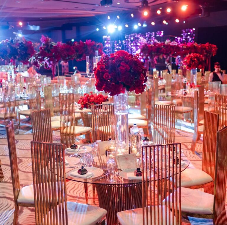 Tables with tall red and pink flower arrangements and gilt chairs, Vivaah Weddings