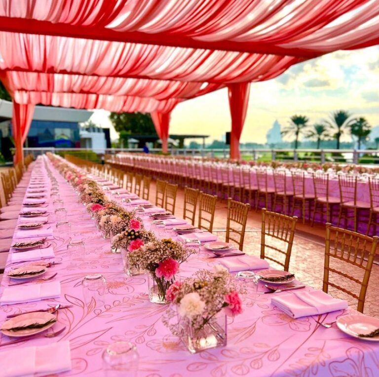 Long pink-clothed table set with small floral arrangements and gold chairs, Vivaah Weddings