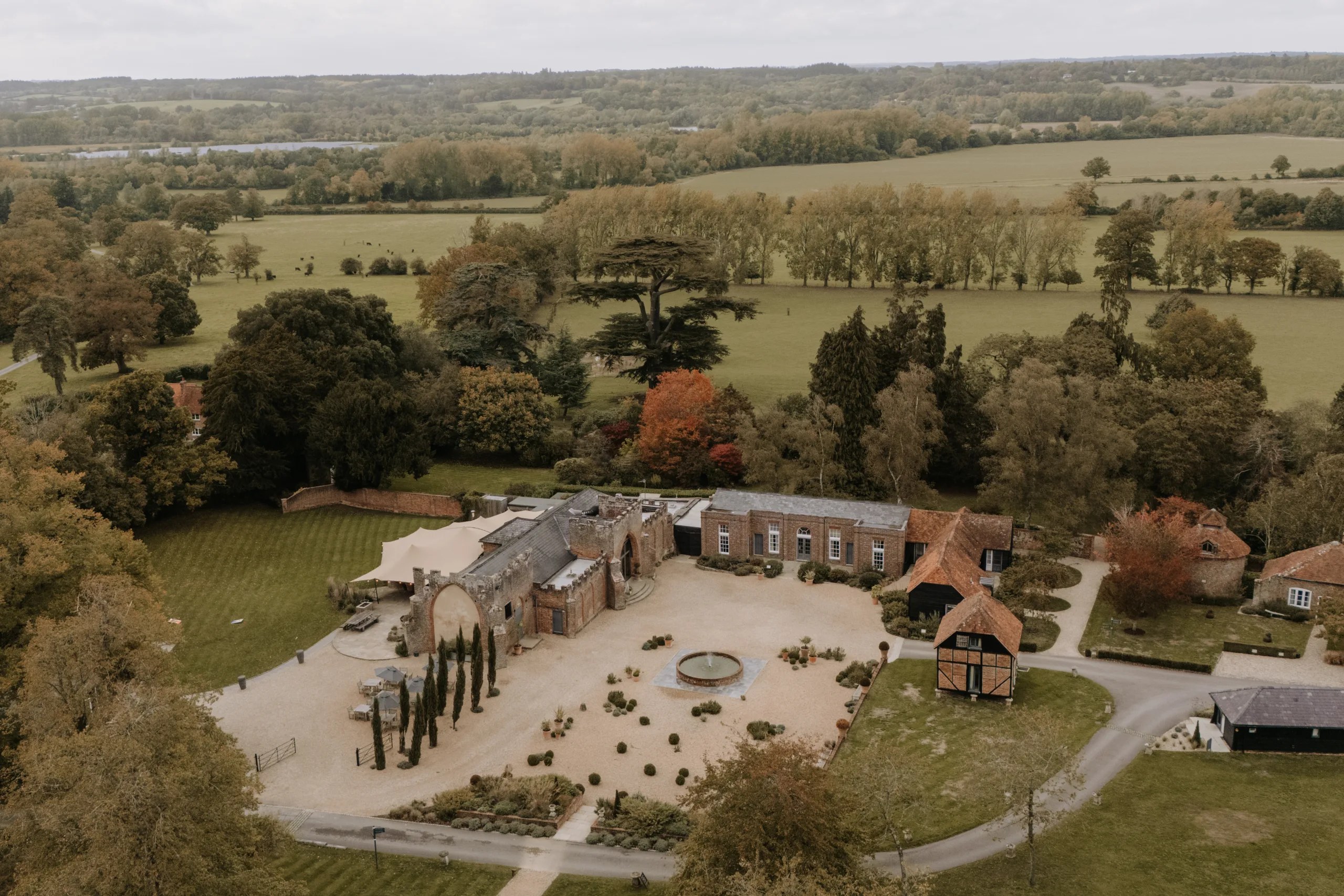 The farm hamlet from the air: the Castle Barn, Coach House and Granary around a gravel courtyard with a fountain, parkland and fields beyond, Wasing Park Weddings