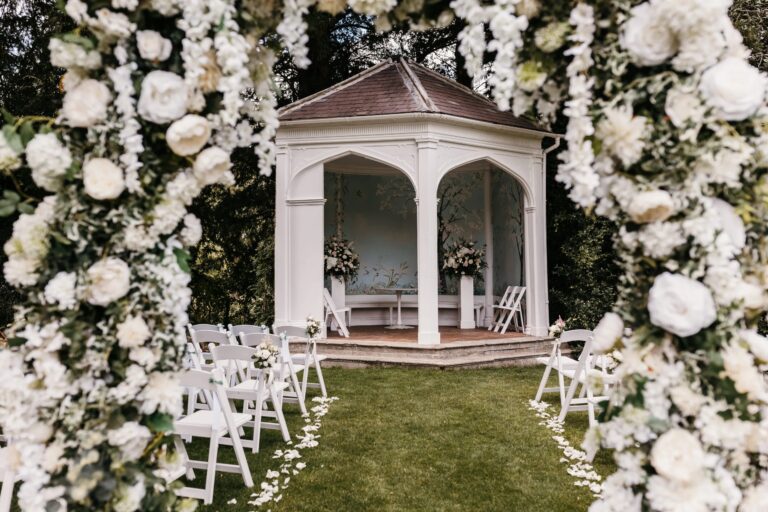 The white Victorian Summerhouse set for an outdoor ceremony, framed by an arch of white flowers with chairs on the lawn, Wasing Park Weddings