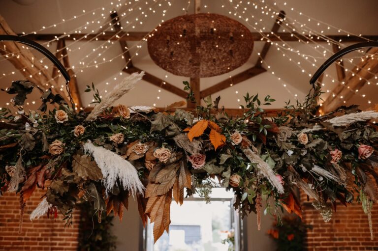A suspended installation of dried flowers, pampas and roses beneath the Castle Barn's lit timber ceiling, Wasing Park Weddings