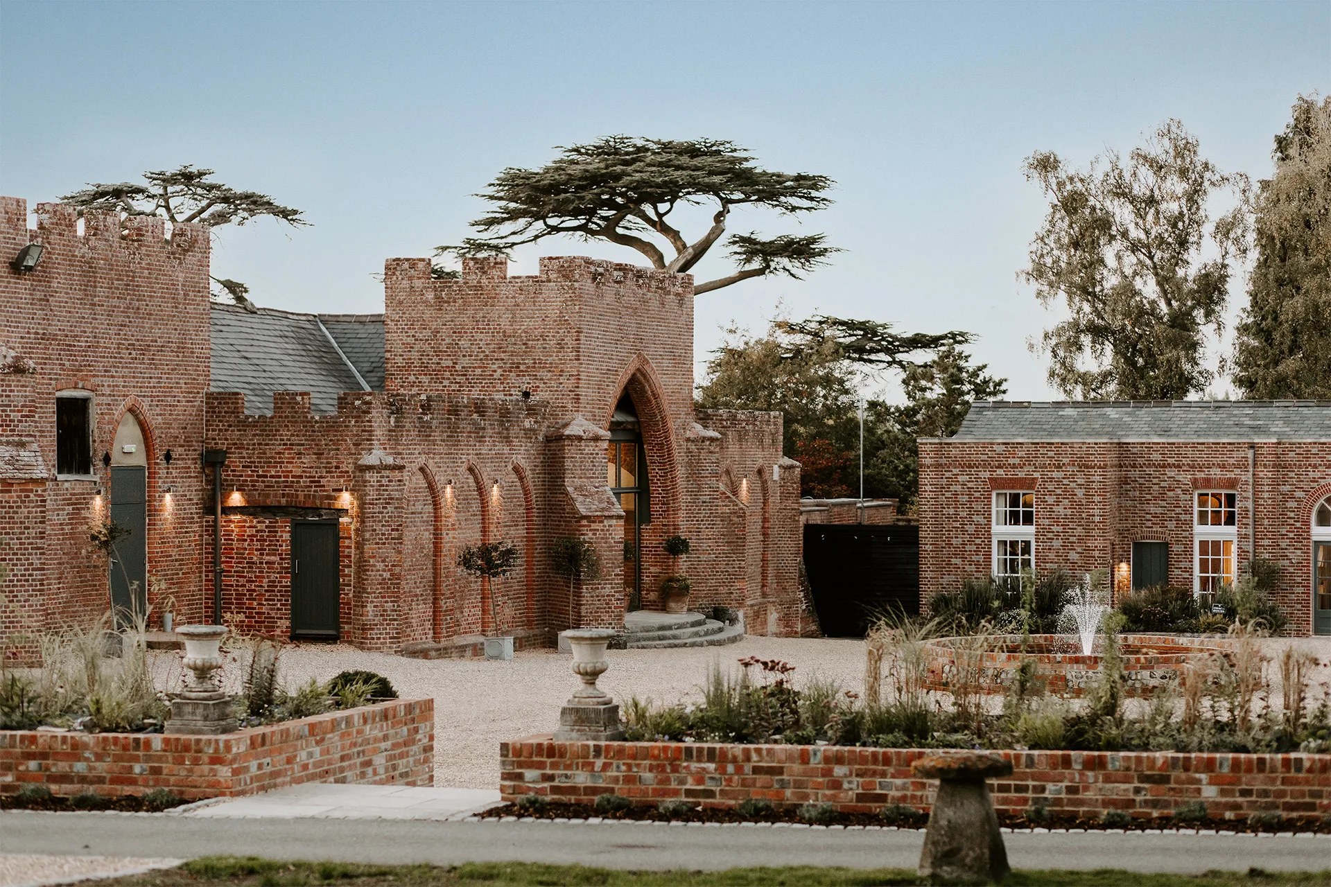 The crenellated brick front of the Castle Barn with its pointed arched doorway, cedars behind and the walled garden beds in front, Wasing Park Weddings