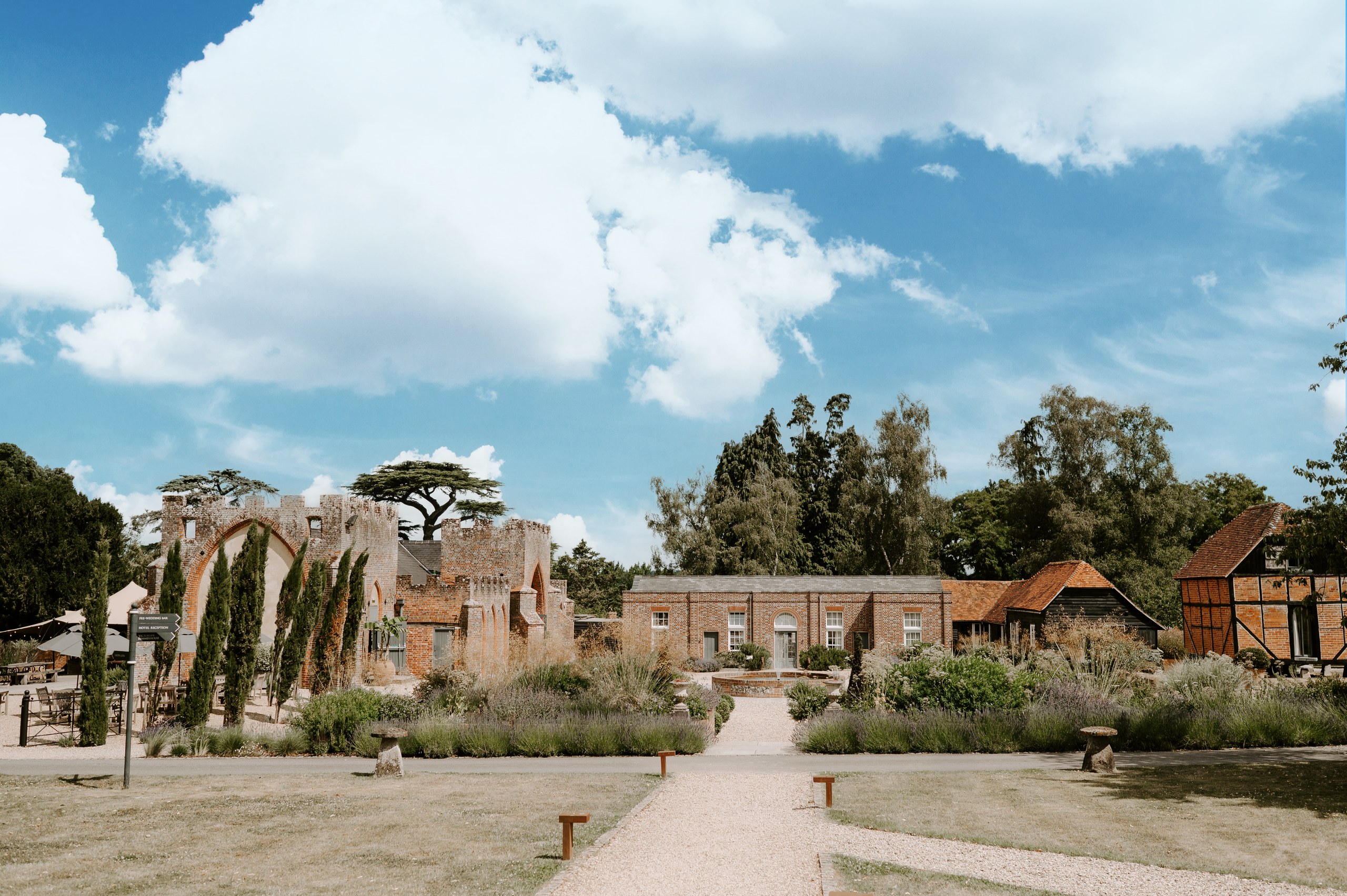 The Castle Barn, Coach House and timber-framed Granary seen across the lawns and gravel paths on a bright day, Wasing Park Weddings