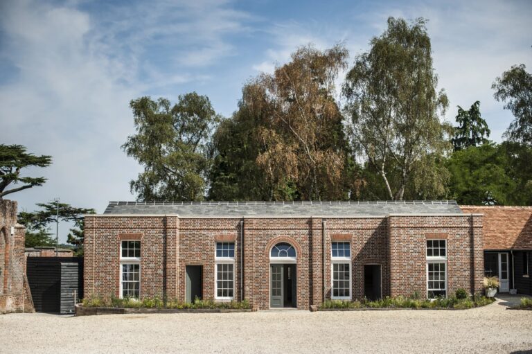 A long red-brick building of guest rooms facing a gravel yard, with tall trees behind, Wasing Park Weddings