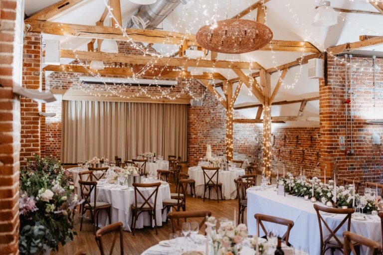 Inside the Castle Barn laid for a wedding breakfast: round tables, cross-back chairs, oak trusses, fairy lights and the stage curtain, Wasing Park Weddings