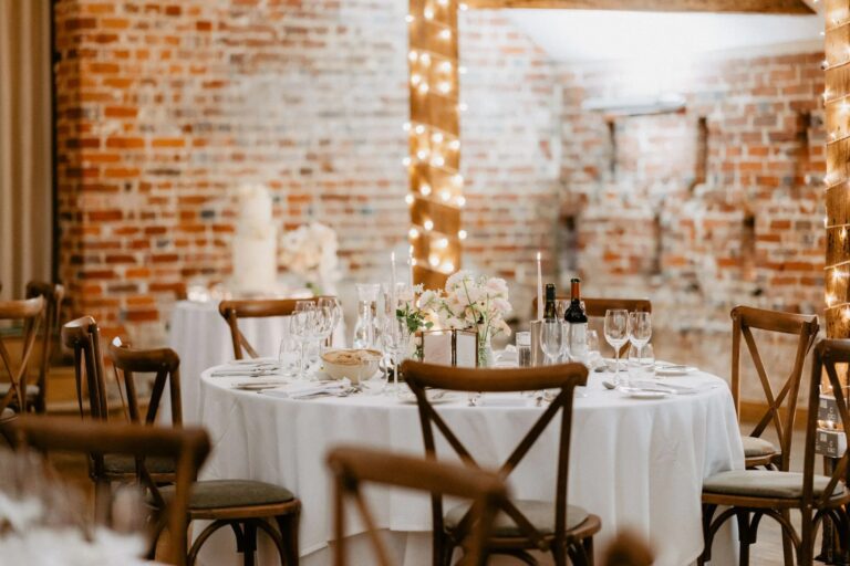A round table dressed in white against the barn's brick walls, with the wedding cake on its own table behind, Wasing Park Weddings