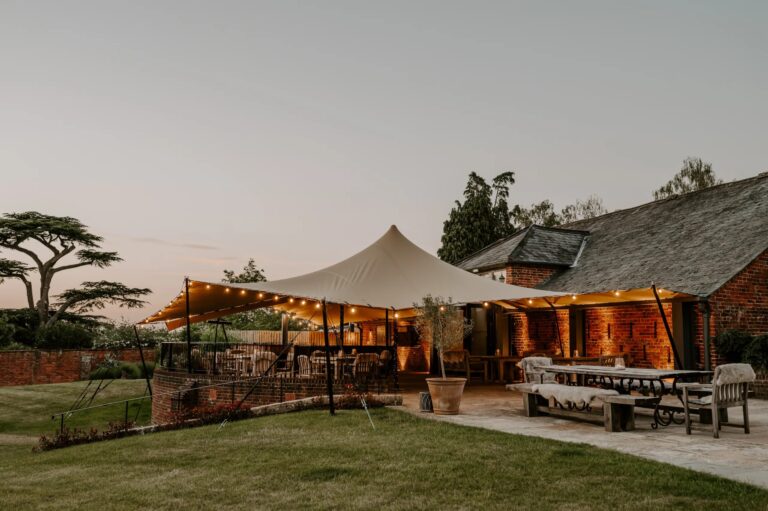 The Castle Barn terrace at dusk under its lit stretch tent, with long tables and benches on the paving, Wasing Park Weddings