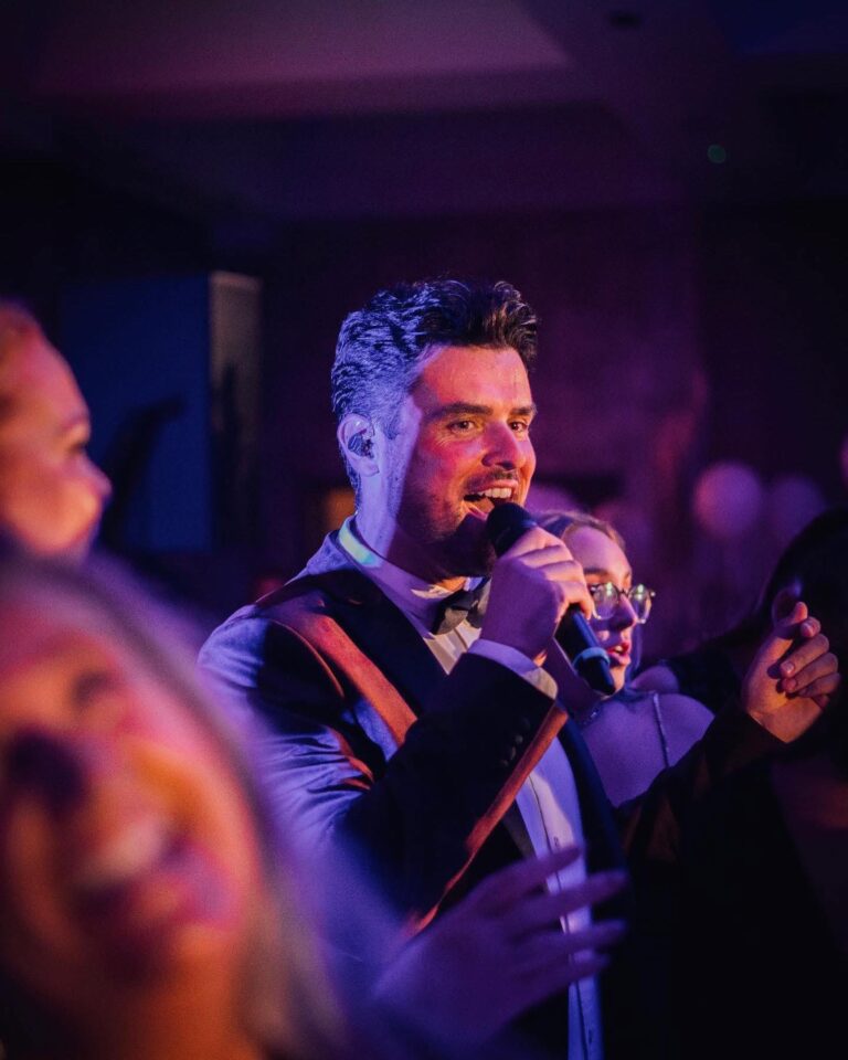 A male vocalist singing among guests in a lit room, Waterfront