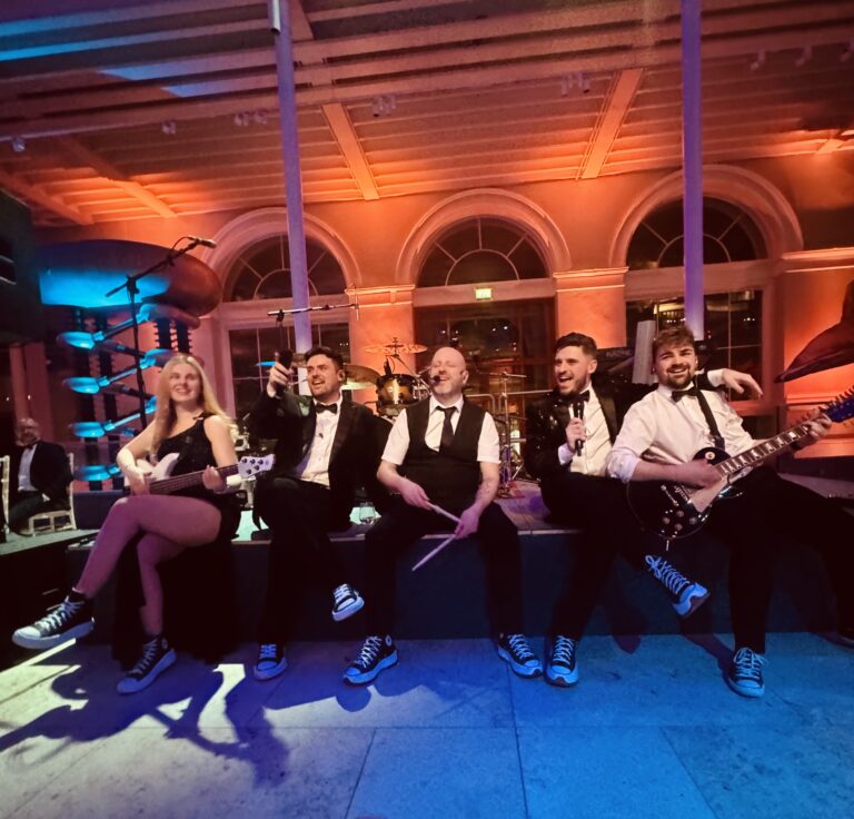 The band seated together on a stage edge in an orange-lit hall, Waterfront