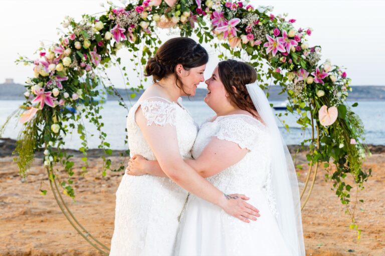 Two brides embracing under a floral arch on the beach, Wed In Malta