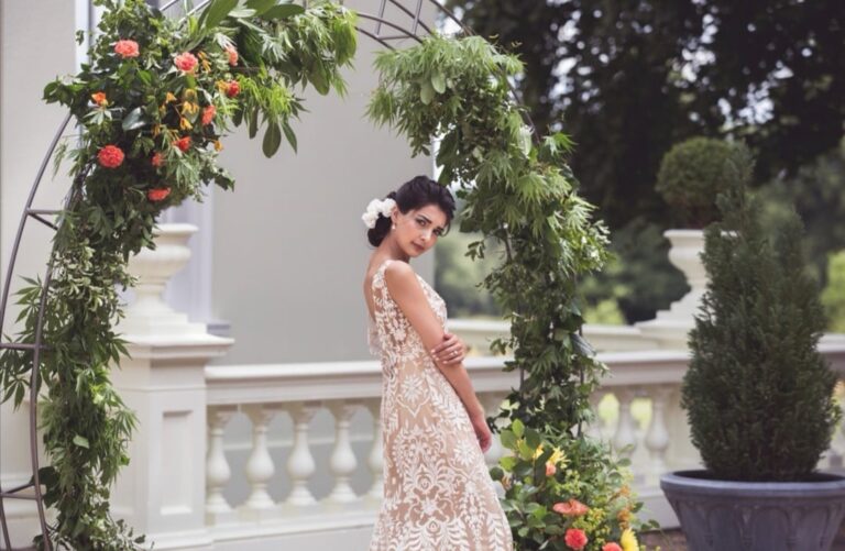 A bride glances back beneath a flower-covered arch, a blush lace gown, Wedding Hair And Makeup By Lisa