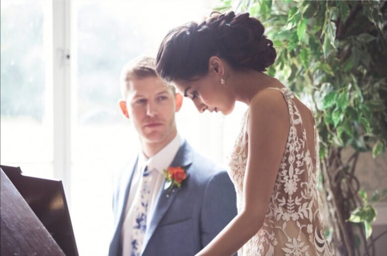 A bride in a low-back lace gown, her groom looking on, indoors among trailing greenery, Wedding Hair And Makeup By Lisa