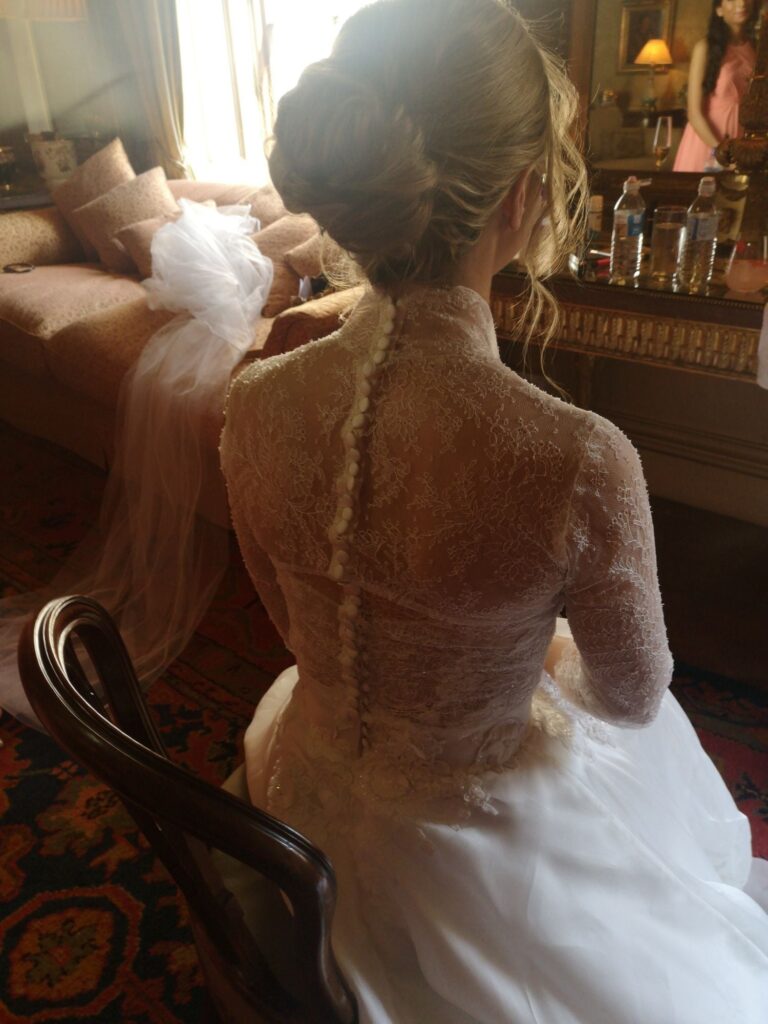 A bride seen from behind, a lace gown with covered buttons, veil beside her, dressing-room window light, Wedding Hair And Makeup By Lisa