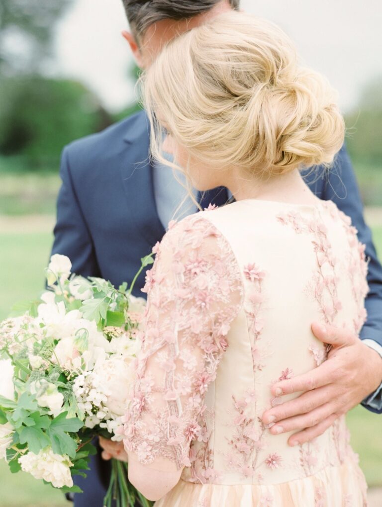 A bride embraces her groom from behind, a blush 3D floral lace gown, a white bouquet, Wedding Hair And Makeup By Lisa