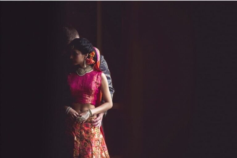 A bride and groom silhouetted in a doorway, her pink and orange sari catching the light, Wedding Hair And Makeup By Lisa