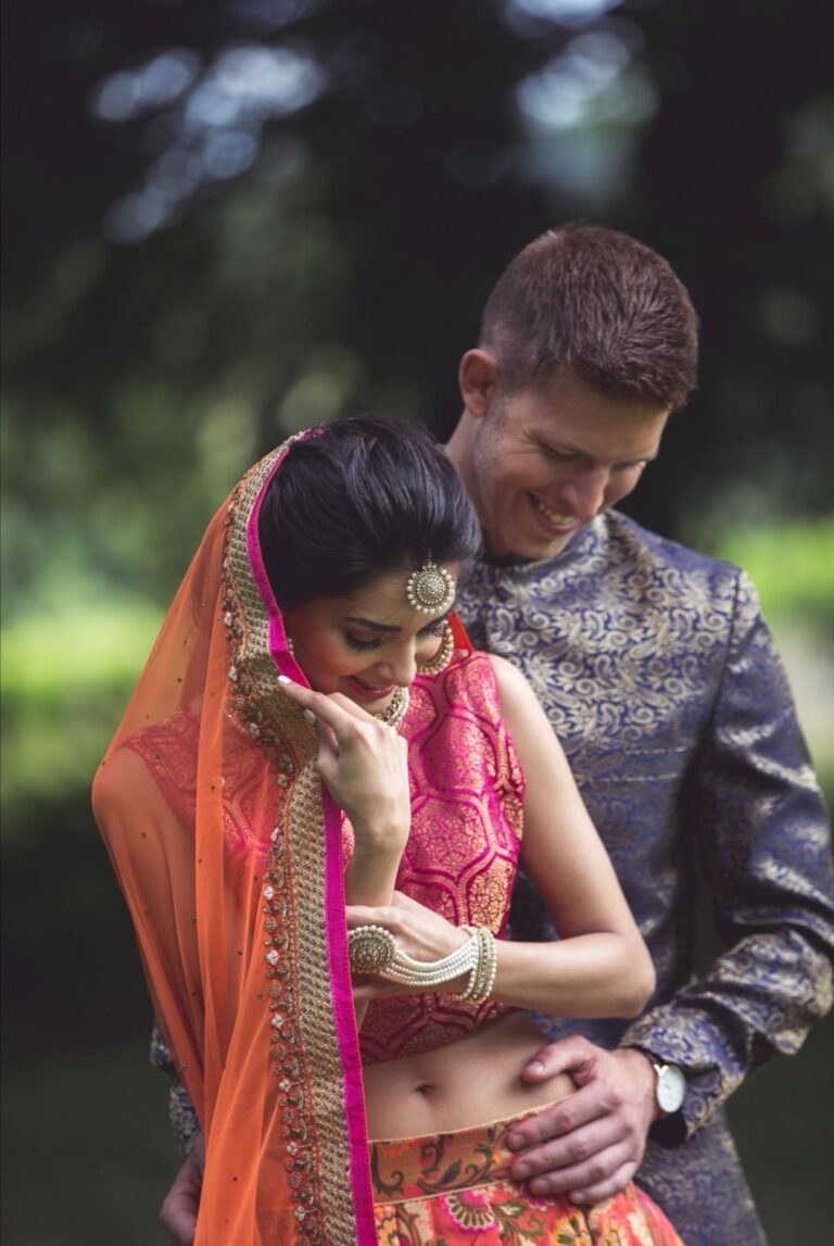 A bride in an orange dupatta laughs with her groom in a navy sherwani, a garden setting, Wedding Hair And Makeup By Lisa