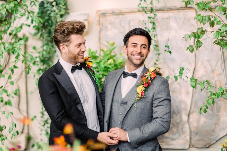 Two grooms hold hands and smile, floral buttonholes, a stone wall behind, Wedding Hair And Makeup By Lisa
