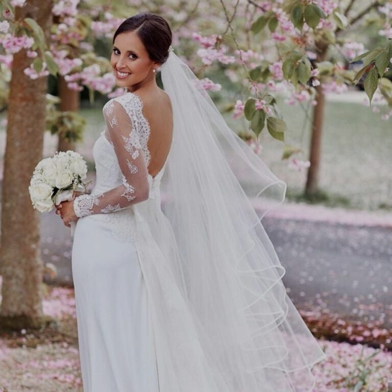 A bride glances back beneath cherry blossom, veil trailing, a white bouquet, Wedding Hair And Makeup By Lisa