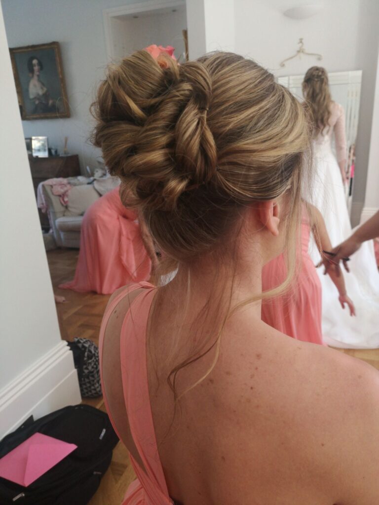 A bridesmaid's twisted updo finished with a pink rose, the bride visible in the mirror behind, Wedding Hair And Makeup By Lisa