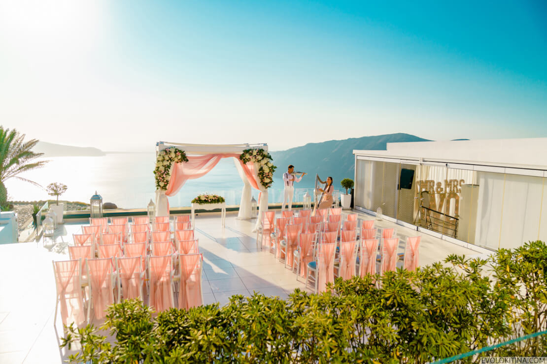 Rows of pink chairs and floral arches for a ceremony on a Santorini terrace, Wedding in Greece