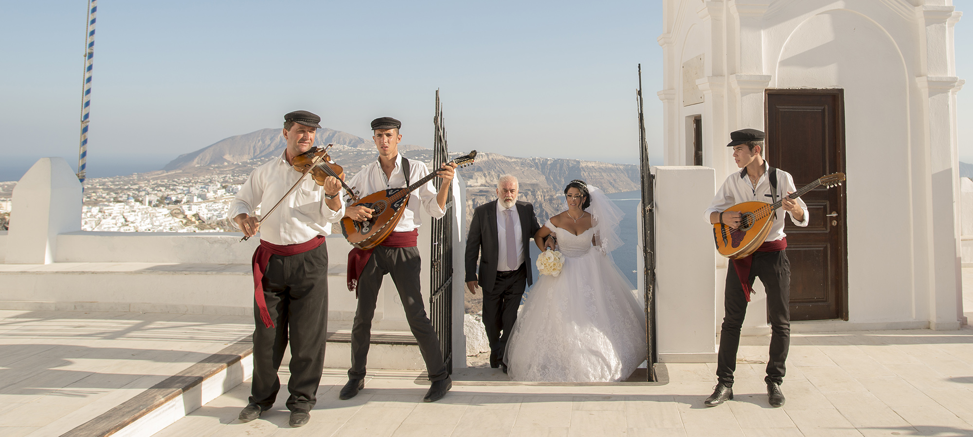 Musicians with guitars lead the bride towards a whitewashed chapel in Santorini, Wedding in Greece