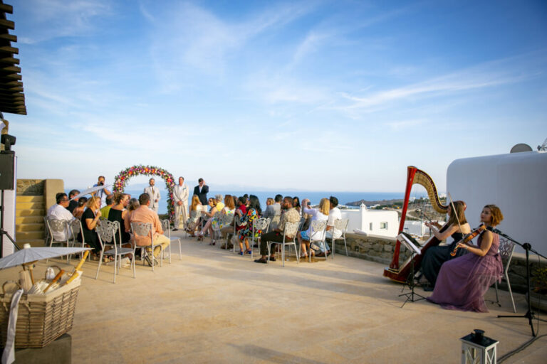 Guests gather beneath a floral arch with a harpist beside the sea, Wedding in Greece
