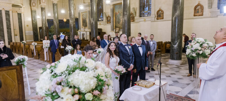 Guests and priest in a church ceremony with large white flower arrangements, Wedding in Greece