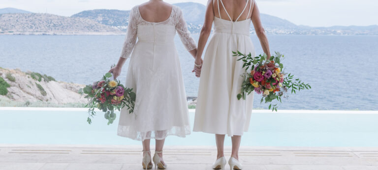 Two brides hold bouquets and look out over a pool and the sea, Wedding in Greece