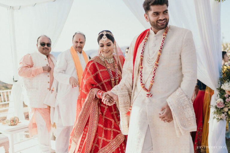 A bride and groom in Indian wedding dress walk with family under a white canopy, Wedding in Greece