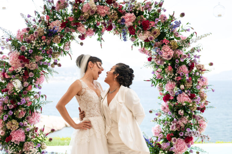 Two brides embrace in front of a round arch of pink and purple flowers, Wedding in Greece