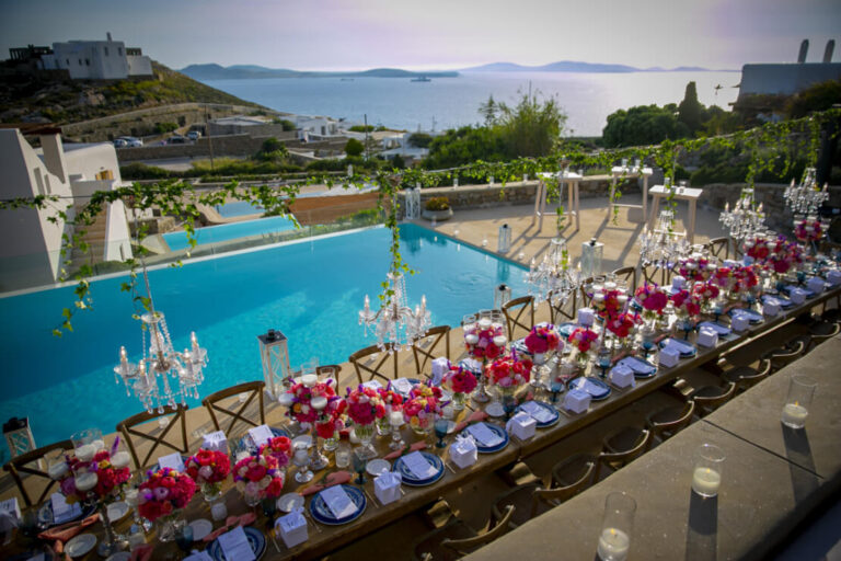 A long reception table with red and pink flowers and chandeliers beside a pool, Wedding in Greece
