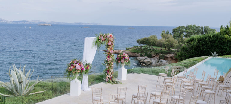 A ceremony set up with a floral arch beside a pool and the sea, Wedding in Greece