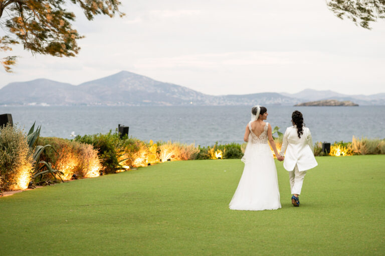 Two brides walk hand in hand across a lawn towards the bay, Wedding in Greece