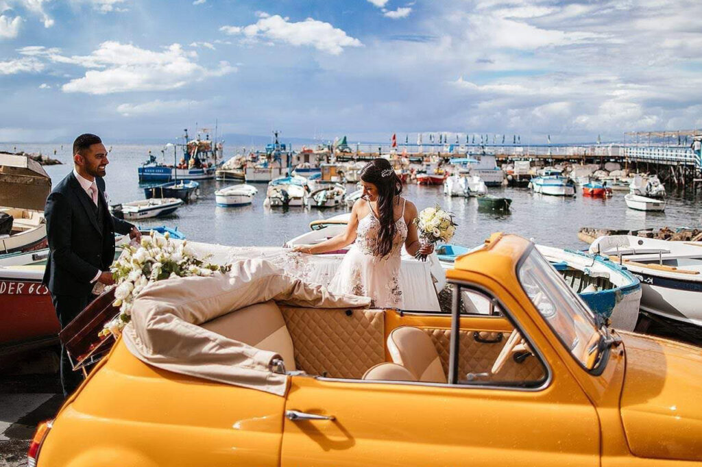 A bride stepping into the Fiat 500 on a harbour quay with fishing boats