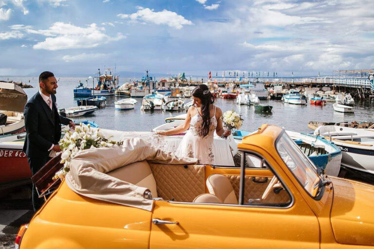 Bride stepping into the Fiat 500 on a harbour quay with fishing boats, Wedding In Yellow Vintage
