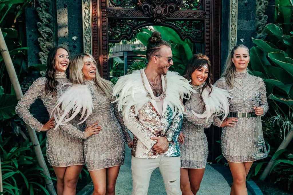 Five band members in sequins and feathers in front of a carved wooden door and tropical planting