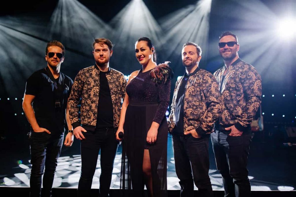 Four men and a woman in patterned jackets on stage in front of stage lights
