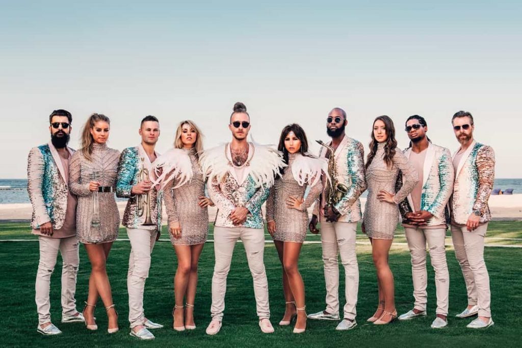 A ten-piece band in white and pastel outfits on a lawn above the sea