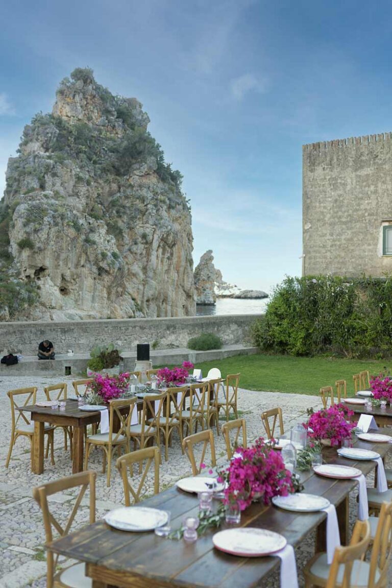 Reception tables with bougainvillea beside a sea stack and an old tower, Wedding Sicily